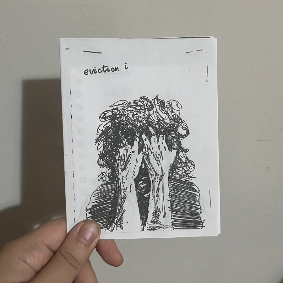 Image of a hand holding up a small paper zine with a black and white ink drawing on the cover